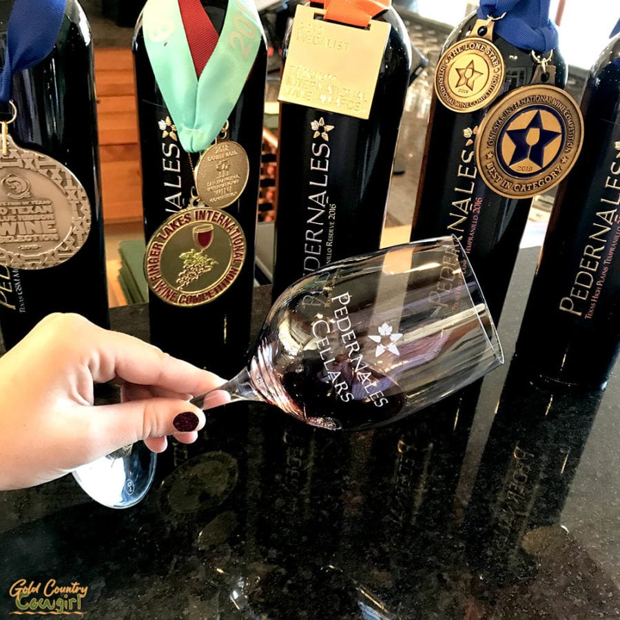 hand holding wine glass in front of bottles with medals at Pedernales Cellars, one of Fredericksburg's best wineries