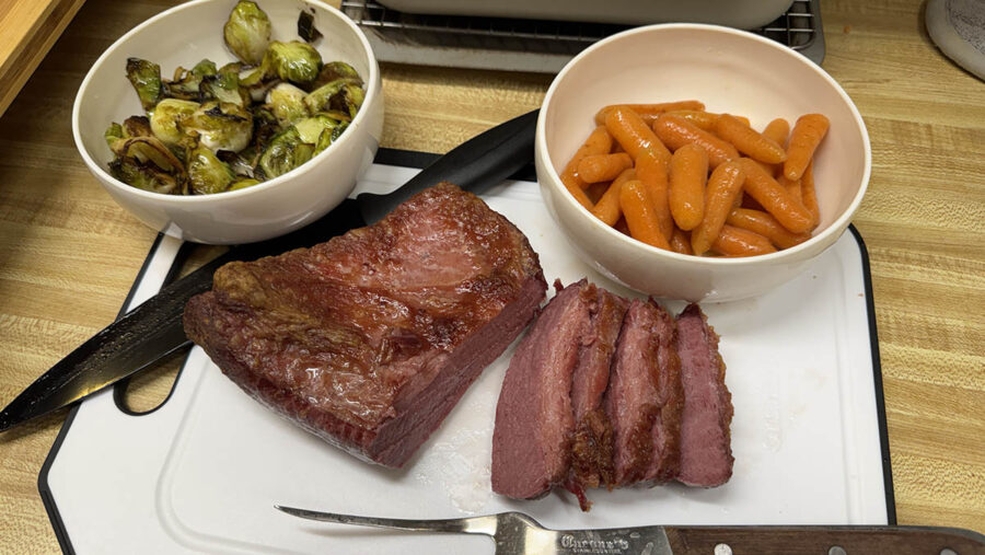 A cutting board with sliced corned been and bowls of carrots and Brussels sprouts